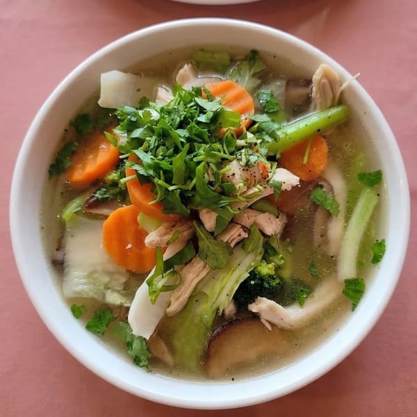 A white bowl of Vietnamese chicken soup with carrots, broccoli, and fresh herbs.
